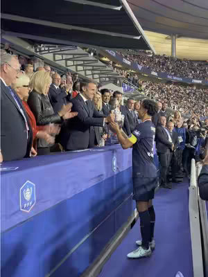 A photograph captures a moment at a stadium, possibly after a match. A soccer player holds up a trophy with others. In the foreground, a blue barrier separates the crowd from the field. People, including those in suits or official attire, stand and applaud. The surroundings are filled with stadium seating. Colors include blues, whites, and shades of grey.
