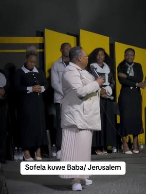 The cover image shows a group of people inside a church or auditorium setting. The foreground features a person in a light-colored coat and skirt, holding a microphone and speaking. Behind them, several other individuals are visible, with some appearing to be part of a choir or worship team. The background consists of yellow panels and a dark wall, which suggest a stage setup. The image conveys a sense of a gospel show or a worship service.