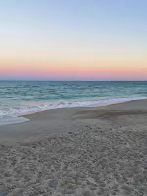 A serene beach scene with a gradient sky. The sky transitions from light blue at the top to pink and orange at the horizon, where it meets the ocean. The sea is a turquoise color, with gentle waves washing onto the sandy beach. The beach is in the foreground, with varying textures of sand visible.