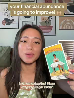 A woman with long dark hair and a white top holding up a tarot card. Above her, text reads 'your financial abundance is going to improve!' in beige. Below her, the caption states, 'but I am seeing that things are going to get better'.