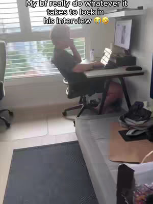 A person is sitting in a room, appearing to do a work interview at a computer, while they are on the phone. The person is in an office-like setting with a window, desk, and monitor.