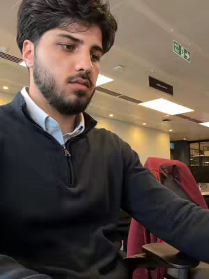 A man with dark hair and a beard is shown in an office setting. He is wearing a dark blue zip-up sweater over a white collared shirt. The background includes office lights, a red chair, and a ceiling.