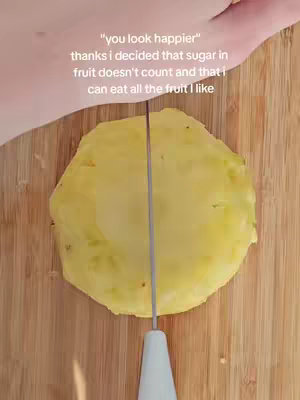 An overhead shot captures a wooden cutting board with a vibrant yellow pineapple slice in the center. A knife is positioned, cutting the pineapple in half. The text overlay adds context, stating that the creator is "happier" and deciding that fruit sugar doesn't count, allowing them to eat as much as they like.