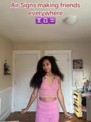 A woman with long dark hair stands in a room with white walls, two closed white doors, and a ceiling light. She is wearing a pink top and pink pants. Text overlay reads: Air Signs making friends everywhere, with Gemini, Libra, and Aquarius zodiac symbols.
