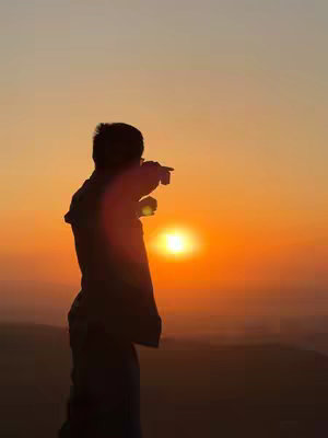 A silhouette of a person standing on a hill, pointing towards the sunset. The sky is a gradient of orange and yellow, with the sun positioned in the center.