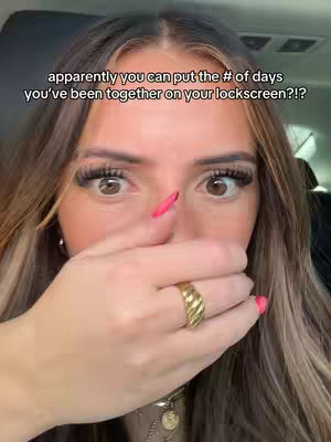 A selfie-style shot of a person with long, flowing hair, angled slightly from a high perspective. The person's right hand covers their mouth. The foreground shows a close-up with emphasized eyes looking in the viewer direction, and a hand covering the lower face and mouth with red nail-polished finger. The background has a blurred car interior.