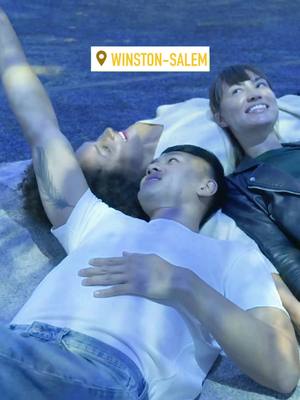The cover image displays three individuals lying down, looking upwards as if gazing at something overhead. The central figure, an Asian male, is the most prominent, with his arms raised and tattoos visible on his left arm. To his right, a Caucasian female smiles and looks upwards, while partially obscured on the left is another person, also looking upwards. The background is a muted blue, and a location pin icon with the text 'Winston-Salem' is overlaid at the top.