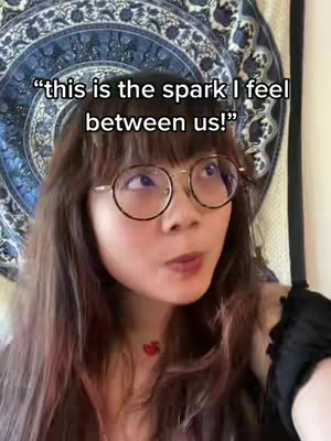 A young woman with long, brown hair and round glasses is looking up with an expression of thought or wonder. She is positioned in front of a patterned tapestry in shades of blue and white. The lighting seems natural, possibly from an indoor setting. The woman is wearing a black top with a contrasting floral accessory near her shoulder. The text overlay indicates an astrology theme for the video's content.