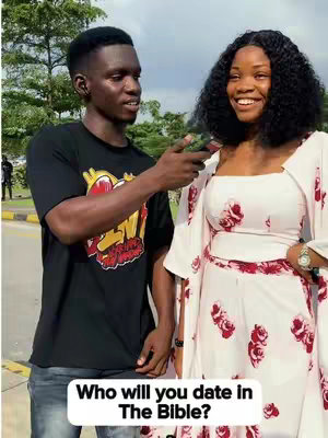 A man interviews a smiling woman on a sunny day. The man is pointing a phone towards the woman, who is wearing a floral outfit. Text in a white box at the bottom of the image asks: 'Who will you date in The Bible?' The background includes trees, pavement, and other people.