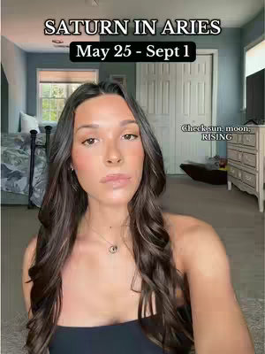 A woman with long, dark hair is centered in the frame, facing the camera. Above her is a text overlay with the title 'SATURN IN ARIES' in large, white, serif font. Beneath this, in a smaller, black text box, it reads 'May 25 - Sept 1'. To the right of her shoulder, in a smaller, white text bubble, it says 'Check sun, moon, RISING'. The background seems to be a bedroom setting, with a bed visible on the left, a closed door in the middle, and a chest of drawers on the right.
