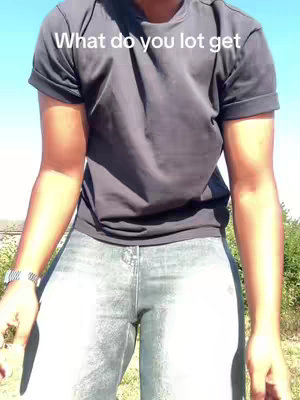 A person stands in front of a blue sky, partially framing the shot from the abdomen up. The individual is dressed in a dark gray t-shirt and light-colored jeans. The text overlay reads "What do you lot get".