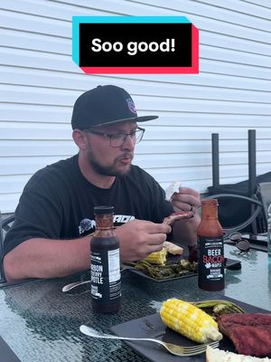 A man in a black baseball cap and glasses is seated outdoors at a patio table. Various BBQ items such as corn on the cob, grilled asparagus, ribs, and bottles of BBQ sauce are on the table. A text overlay that says “Soo good!” is at the top. The background appears to be a white, textured wall.