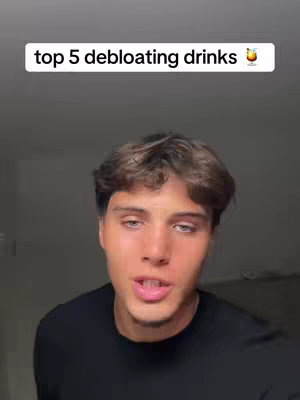 A selfie-style video cover image featuring a young man with brown hair and a black shirt, positioned in front of a plain, neutral grey background. The man's face commands attention, and the focus is on him. A white text overlay at the top reads "top 5 debloating drinks" with a small, stylized glass of a drink emoji right next to it. The color scheme is natural with the man's skin tone and hair color being the most prominent, with the black attire on the other hand providing contrast to the overall composition.