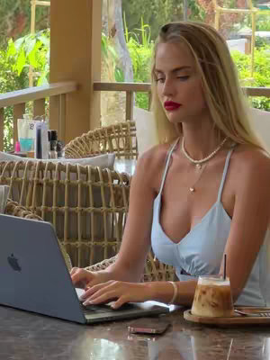 A blonde woman with red lipstick works on a laptop at a cafe. She wears a light blue dress and a pearl necklace. An iced coffee sits on the table in front of her.