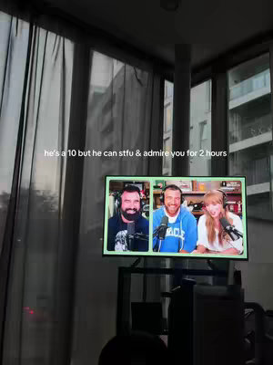 A dimly lit interior shot with sheer curtains in the background. A TV screen displays a podcast with three individuals. Text overlay at the top. The overall composition has a casual, indoor feel.