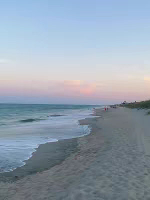 A scenic view of a beach at dusk or dawn, with a vast expanse of ocean meeting a sandy shore. The sky transitions from a soft blue at the top to a pale pink and purple hue towards the horizon, suggesting a sunrise or sunset. The ocean has gentle waves that create a white foam pattern along the water's edge. The sand is a light tan color, and there are possible figures scattered along the beach, near the water's edge.