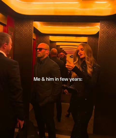 The image showcases an elevator interior with a warm, amber lighting scheme. The walls are clad in a repeating, ornate design, with reflective surfaces that suggest mirrored panels. In the foreground, a woman takes a selfie in front of a man with a bald head and sunglasses. The woman is wearing a black, sequined outfit. The man next to her is wearing a black coat. Reflecting in the mirrored walls are other people. The image suggests a candid moment within a celebrity setting.