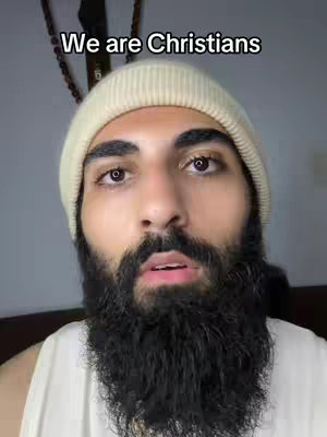 A man with a beige beanie, a large dark beard, and a light sleeveless shirt. He is looking directly at the camera, with text overlaying the image.