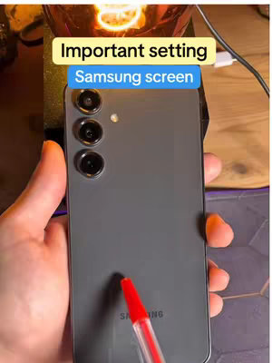 A person's hands holding a grey Samsung phone with the camera lenses visible. A pen is pointing towards the phone. Text overlays the image: 'Important setting' and 'Samsung screen'. The background is dimly lit, with lights creating warm tones.