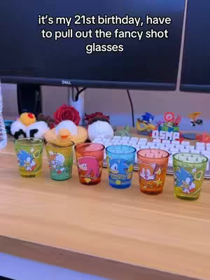 A collection of Sonic the Hedgehog themed shot glasses are displayed on a wooden surface. In the background, there are computer monitors and other decorative items. Text overlayed at the top of the frame says: 'it's my 21st birthday, have to pull out the fancy shot glasses'.