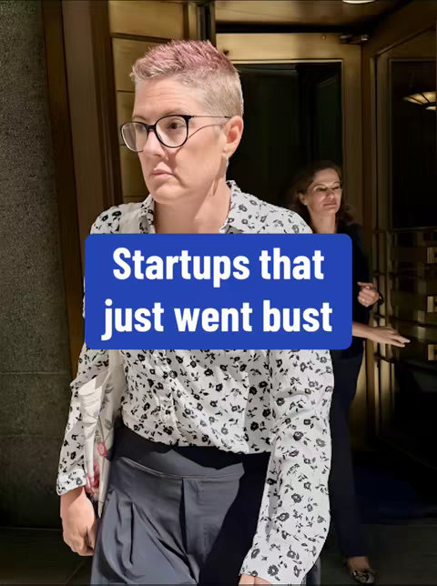 The image captures a woman with short, pink-tinted hair, wearing glasses and a patterned blouse, walking out of a building. A blue banner at the bottom reads "Startups that just went bust." Another woman is partially visible in the background, also seemingly exiting the building. The setting includes a brown door and gold accents.