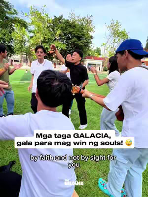 A group of young adults gathers outdoors on a sunny day. They are arranged in a loose circle on a grassy area, with some holding instruments and others singing or gesturing. The composition is dynamic and captures a sense of community and shared activity. The primary colors are greens and blues from the setting, with contrasting white shirts.