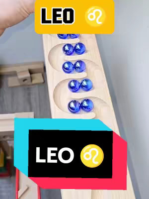 The image shows a wooden marble run with blue glass marbles in a flowing pattern. Overlaid at the top is a yellow rectangle with the word "LEO" in bold, black text and a Leo zodiac symbol. A second rectangular overlay at the bottom repeats the "LEO" and zodiac symbol, with a black background and a pink and blue border.