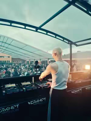 A person is DJing at an outdoor music event, facing away from the camera towards the crowd. The person is wearing a white tank top, black pants with white stripes down the side, and has short, light-colored hair. The DJ setup is prominently displayed, with the crowd visible in the background under a tent. The brand name 'Verknipt' is displayed.