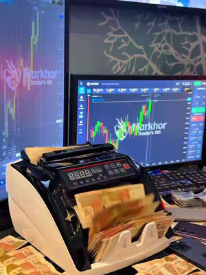 An image showing a money counting machine with green currencies inside. There are two computer monitors in the background that show line graphs. Multiple stacks of currency are visible in the foreground on the table.