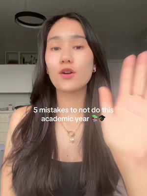 A young woman with long dark hair is holding her hand up towards the camera. Overlayed text says, "5 mistakes to not do this academic year" with book and graduation cap emojis. The background is out of focus, with a light fixture visible behind the subject.