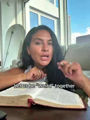 A woman with long dark hair is speaking to the camera. She has visible makeup and is wearing a white top. The background includes a window with a city view, and a portion of a white screen. The woman is holding a Bible and gesturing with her hands. The hook text "Let us be \"weird\" together" is displayed at the bottom of the frame.