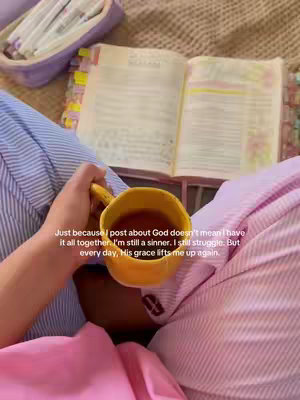 An overhead shot shows a person seated with a Bible open on their lap, a hand holding a yellow mug with a brown liquid. The person is wearing pink clothing and is sitting on a beige surface. In the background, a purple pouch with white pens is visible. The image has a warm, inviting tone.