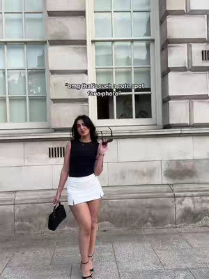 A woman stands in front of a building with large windows, wearing a black top, white skirt, and heels. She holds sunglasses and a purse.
