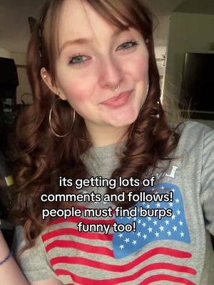 A young woman with reddish-brown curly hair, hoop earrings, a nose ring, and a grey graphic t-shirt with a distressed American flag design is smiling at the camera. Text overlay indicates content about finding burps funny.