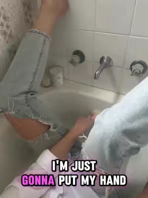 A selfie-style shot taken in a bathroom. The subject's legs are visible, clad in ripped jeans, resting inside a white bathtub. The upper body, dressed in a white shirt, is partially seen, and a hand is reaching towards the water. Text overlays featuring white and pink fonts highlight a humorous statement, suggesting a casual and potentially relatable action.