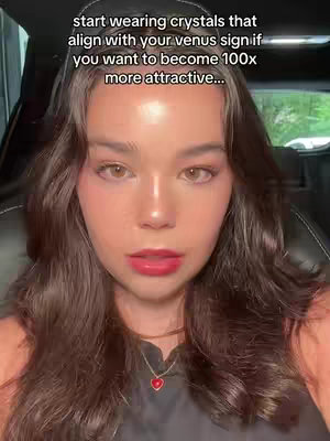 A selfie-style shot featuring a woman with long, dark hair, gazing towards the camera. She has light skin and is wearing makeup, including pink lipstick. The background is blurred, suggesting she's inside a car or similar vehicle.