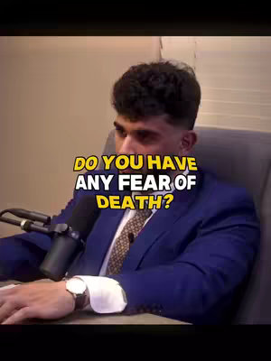 A shot of a man in a blue suit, seated in a chair with a microphone in front of him. The background is a plain, neutral color. The image includes a text overlay.