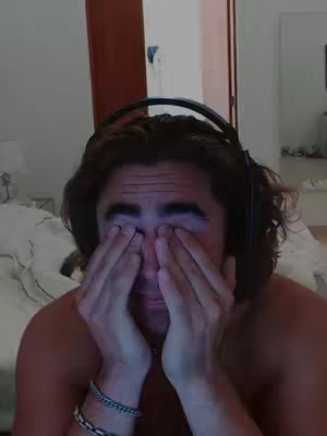 A selfie-style shot of a man with his eyes closed and hands covering them. He has short, tousled brown hair and is wearing a dark headset around his neck. He is shirtless, and visible are the bracelets and necklaces he wears. The background suggests a bedroom setting, with parts of a bed and a doorframe in view, and the entire appearance is lit by soft indoor lighting.