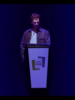 A man stands behind a podium with the words "Letters Live" on it, in front of a dark blue backdrop. The man is wearing a brown jacket over a light blue shirt, the pose suggests a serious or reflective mood.