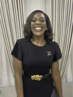 A woman in a black dress with short sleeves, a black belt, and a gold buckle is smiling broadly. She's wearing earrings and a brooch on her dress.