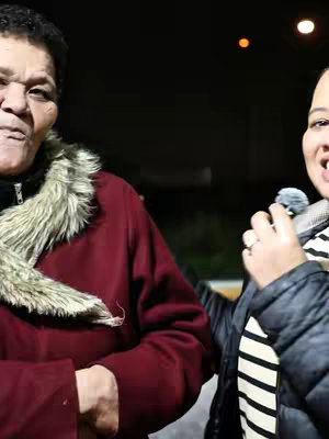 A woman in a striped shirt holds a microphone and smiles, facing the camera at a medium angle from the right side of the frame. An older woman in a red coat is positioned on the left, with a fur collar, looking slightly towards the camera and likely in conversation. The setting appears to be outdoors at night, with a dark background and a street lamp adding warm light to the top right corner.