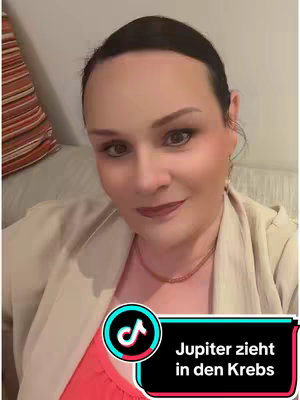 A selfie-style video cover image of a woman with dark hair, fair skin, and makeup. She's positioned in front of a soft, light-colored background, possibly a wall or furniture. The background has a subtle striped pattern. The woman wears a beige blazer over a salmon-colored top and a delicate necklace. She appears to be in an indoor setting, possibly a home. A graphic overlay with the TikTok logo and text is featured prominently at the bottom. The image has a warm, inviting tone.