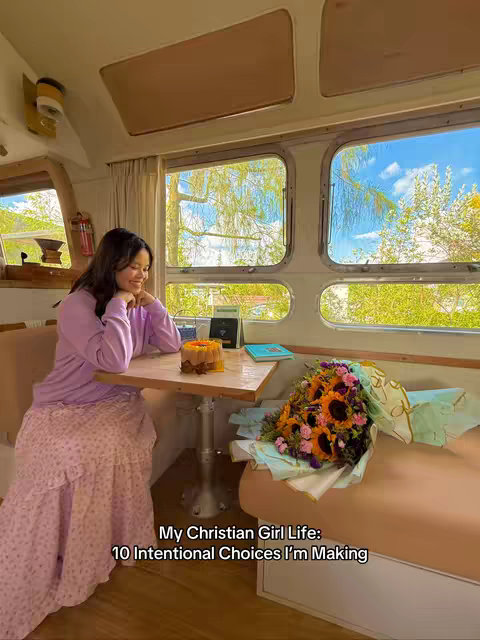 A young woman sits at a wooden table inside what appears to be a converted camper or trailer. She's wearing a purple sweater and a long pink skirt with a floral pattern. A bouquet of sunflowers and other flowers sits on a bench behind her, and a small cake is on the table in front of her. The background includes windows with a view to the outside.