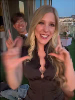 A woman with long blonde hair is smiling, making peace signs with both hands. Another person is slightly visible in the background.