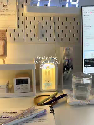 A well-lit study desk setup with a white pegboard, a desk lamp shaped like a bunny with a yellow glow, pens, a digital clock, and a glass of water. The desk also has a notebook, pens, and other stationery items.