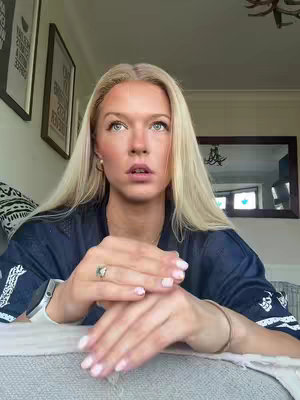 A blonde woman is centered in the frame, looking upwards with a somewhat pensive expression. Her hands are clasped in front of her, with her fingers intertwined. She is wearing a dark blue top, and the background is blurred, showing a glimpse of a room with wall art and a mirror. The lighting suggests it is daytime.