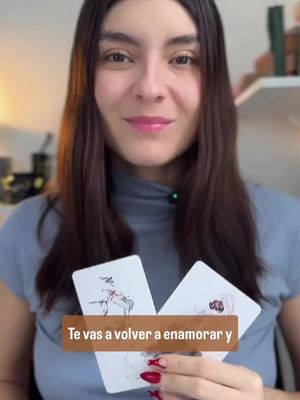 A woman with long brown hair smiles at the camera, holding two tarot cards.  A text overlay at the bottom states, "Te vas a volver a enamorar y". The background is a soft-focus interior.