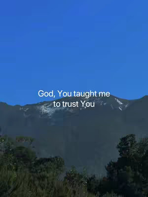 The image shows a scenic view with a clear blue sky above and a mountain range in the background. Dark green bushes cover the foreground. In the center, white text overlaid on top says, 'God, You taught me to trust You'.