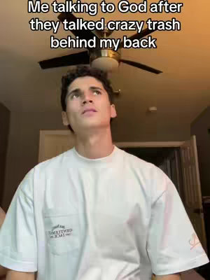 A young man is looking up with a slightly exasperated or thoughtful expression in a bedroom setting. The upper portion of his body is visible, with a white t-shirt that has a pocket with writing on it. The background includes a ceiling fan and a partial view of a doorframe.