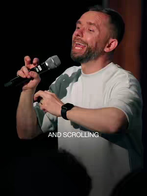 A man with a beard is holding a microphone to his mouth and opening his mouth while standing, with text written next to him. The background is dark and out of focus. The man is wearing a white T-shirt, with a black smartwatch.
